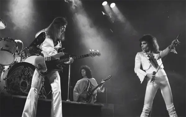 Fotografía en blanco y negro de Queen actuando en un escenario iluminado en 1975